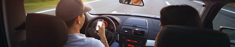View from the back seat of a car: the driver in a cap holds a phone while steering on a road.