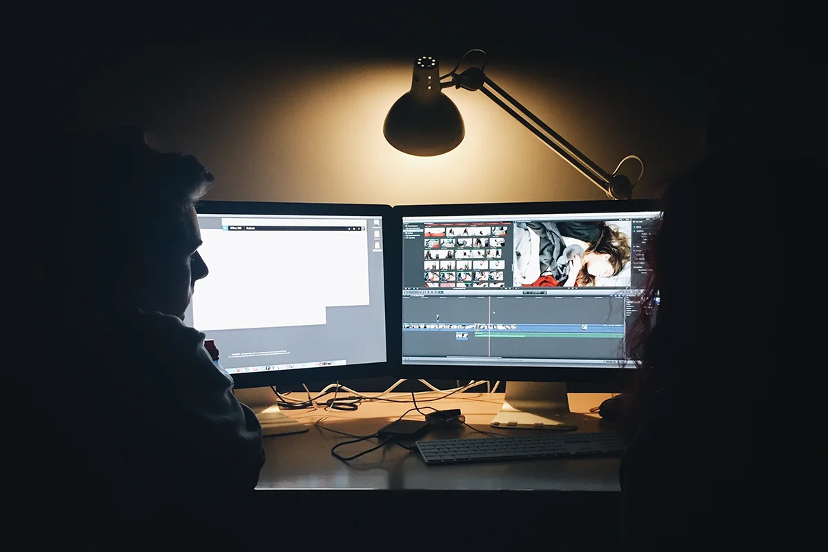 Two people seen in silhouette at a desk, watching video editing software on dual monitors under a lamp.