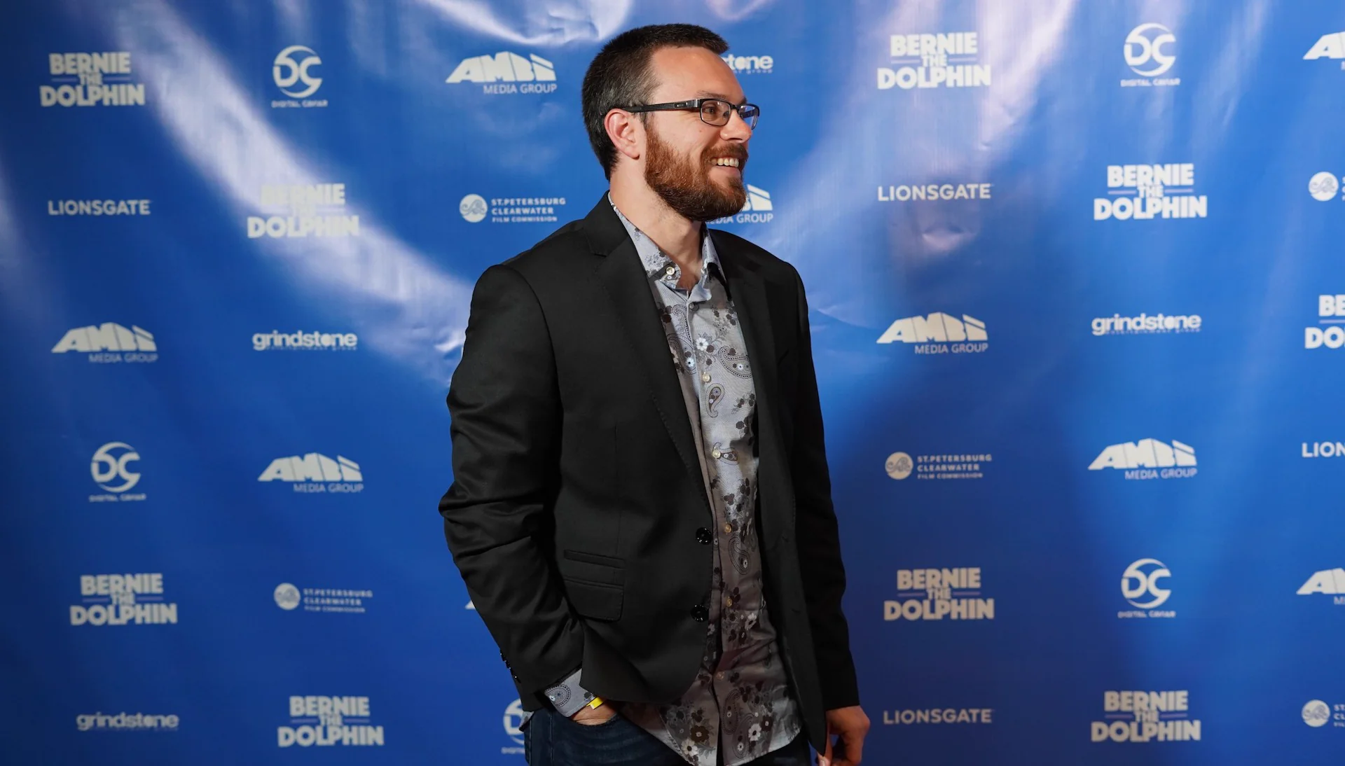 Smiling bearded man in glasses, dark blazer and patterned shirt, standing in front of a blue film premiere backdrop.