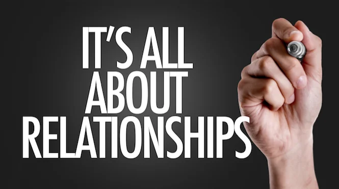 A hand holding a marker beside large white text reading "It's all about relationships" on a dark background.