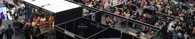 Overhead view of a crowded trade show floor with visitors browsing black-walled booths, including a makeup display.