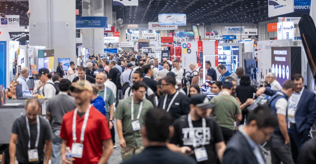 Crowded trade show hall with attendees wearing lanyards walking between exhibitor booths under hanging signs.