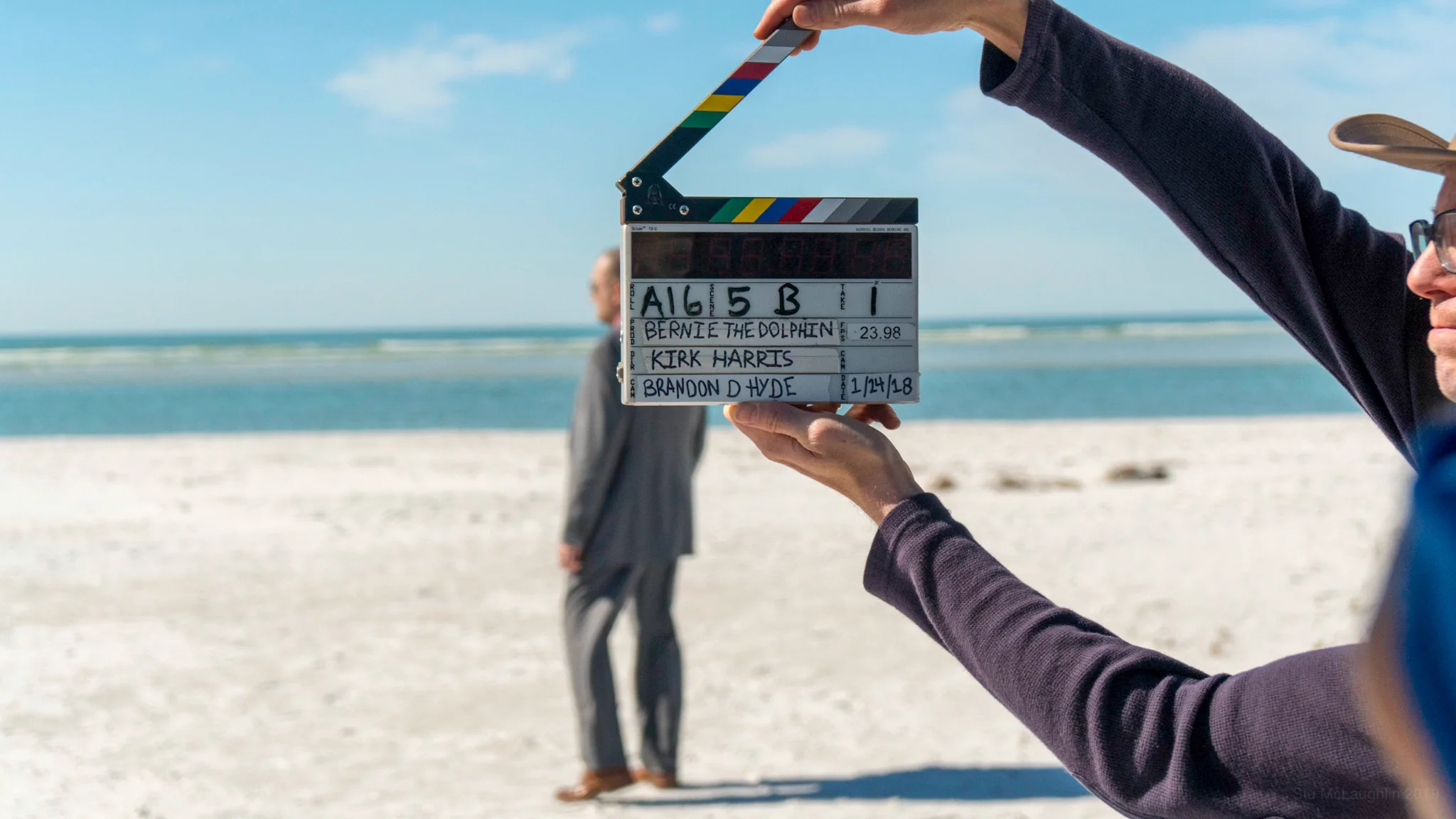 Hands hold up a film clapperboard on a sunny beach, with a man in a grey suit standing blurred behind it.