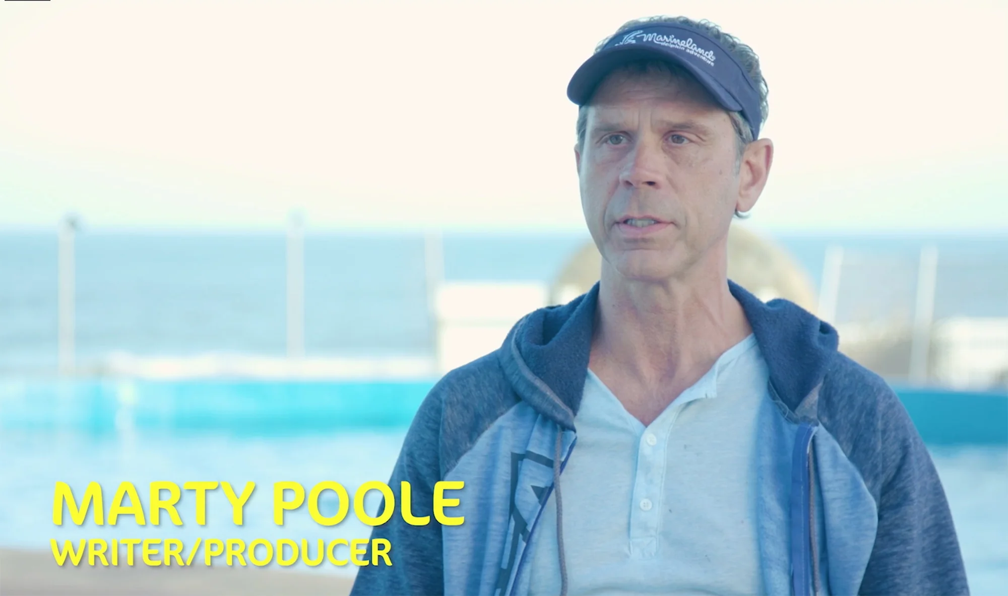 Man in a navy visor and hooded jacket speaks outdoors by a pool and the sea; caption reads Marty Poole, Writer/Producer.