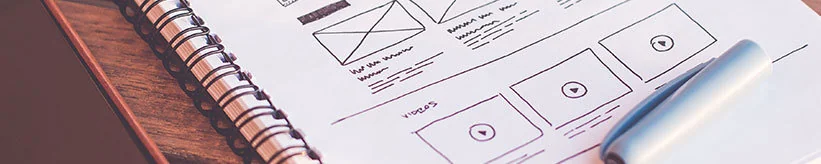 Hand-drawn website wireframe sketches with video play buttons in a spiral notebook, with a silver pen resting on the page.
