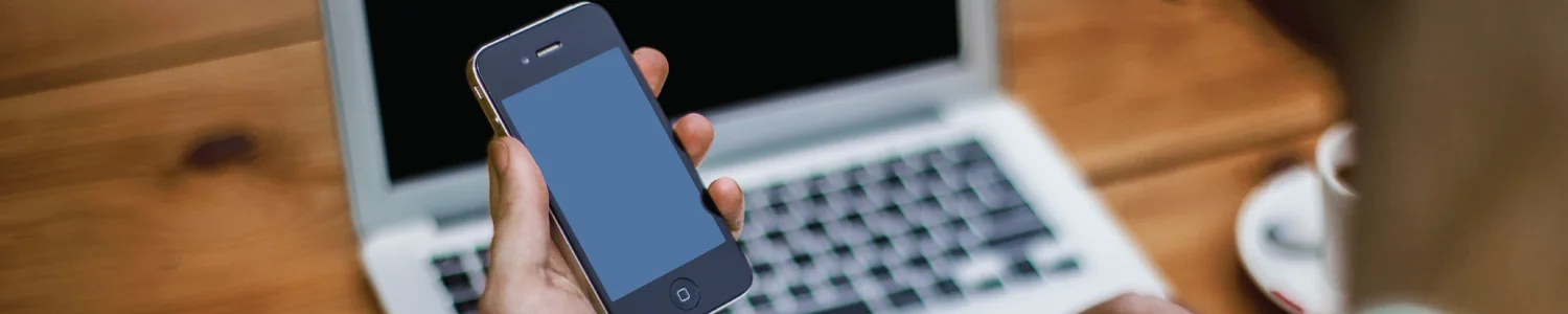 A hand holds a smartphone with a blank blue screen in front of an open laptop on a wooden desk.