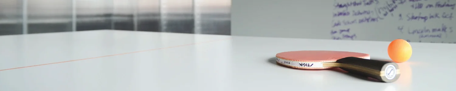 A table tennis paddle and orange ball rest on a white table, with handwritten notes on a whiteboard behind.